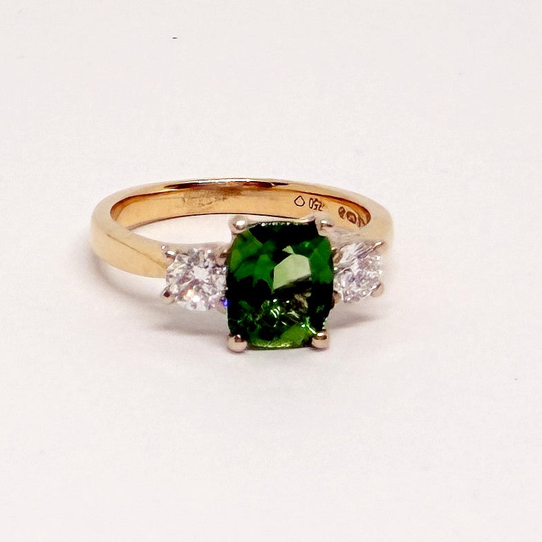 A yellow gold ring featuring a cushion-cut green gemstone in the center, flanked by two small clear round-cut stones on a plain white background.
