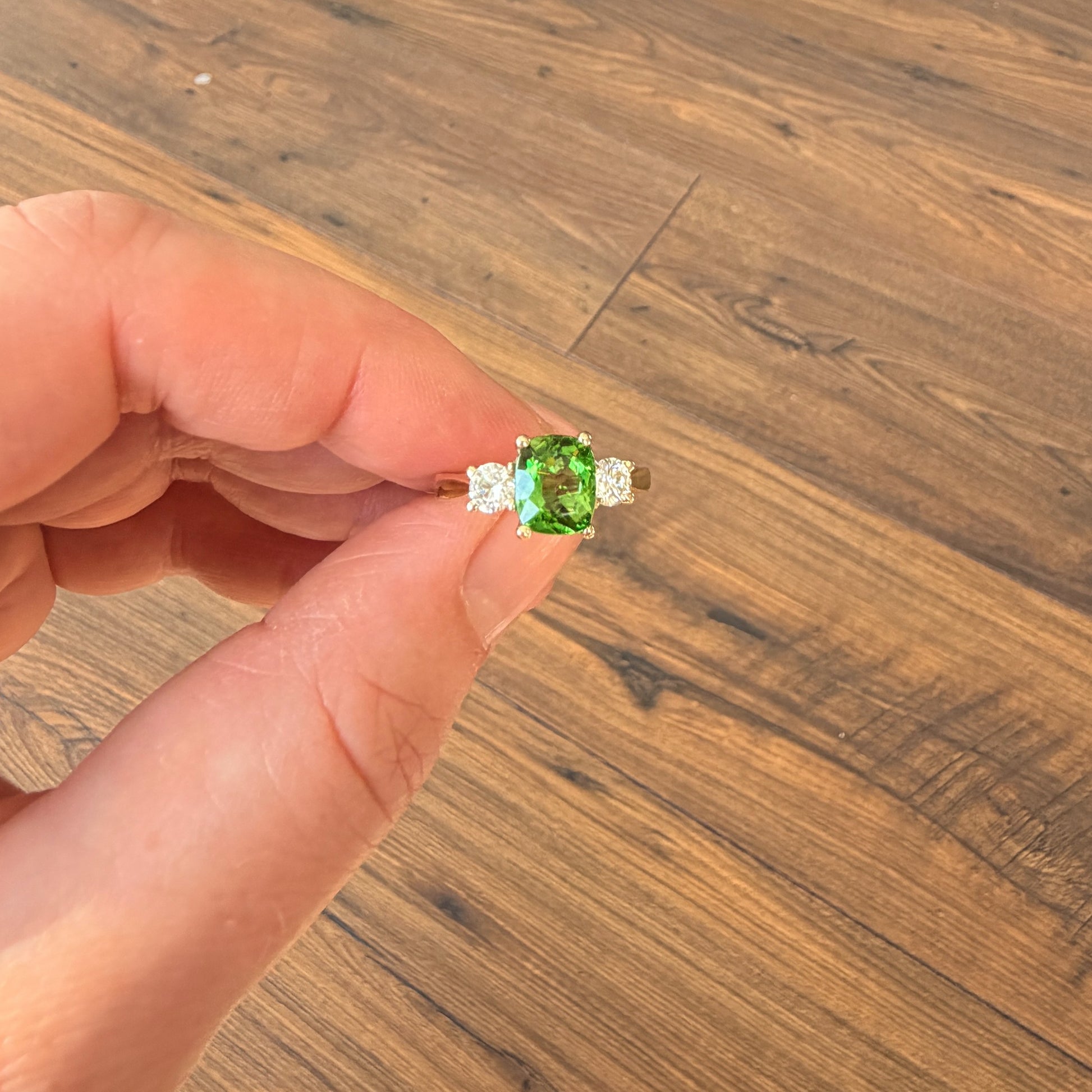 A hand holding a gold ring with a green rectangular center stone and small clear side stones, set against a wooden floor background.